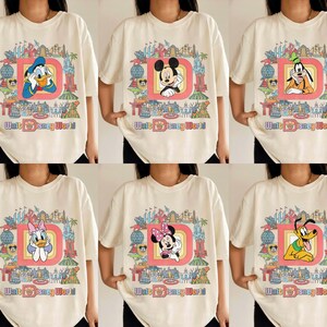 May include: Cream-colored t-shirts featuring various Disney characters inside a red-outlined letter "D" with "Walt Disney World" text. Characters include Donald Duck, Mickey Mouse, Goofy, Daisy Duck, Minnie Mouse, and Pluto. The shirts have a vintage, cartoon style.