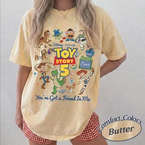 May include: A pale yellow short-sleeve t-shirt featuring a colorful graphic print of Toy Story characters including Woody, Buzz Lightyear, and Rex. The text reads Disney Pixar Toy Story 5, Name, and You've Got a Friend In Me in blue and red.