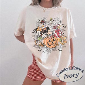 May include: An ivory t-shirt featuring a graphic print of Disney characters in Halloween costumes, including Mickey Mouse, Minnie Mouse, Goofy, Donald Duck, Daisy Duck, and Pluto, with a large jack-o'-lantern and small bats.