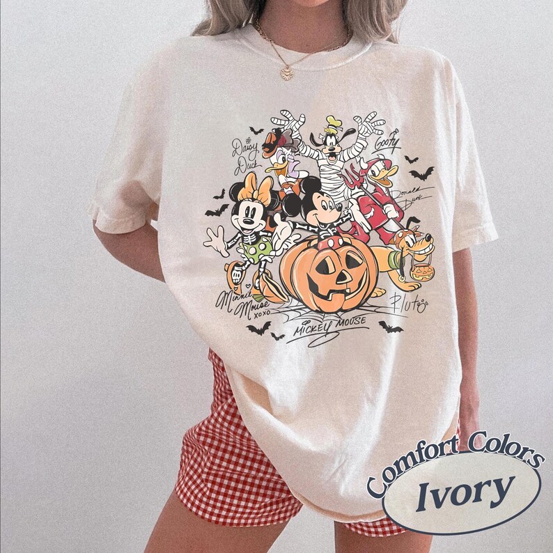 May include: An ivory t-shirt featuring a graphic print of Disney characters in Halloween costumes, including Mickey Mouse, Minnie Mouse, Goofy, Donald Duck, Daisy Duck, and Pluto, with a large jack-o'-lantern and small bats.