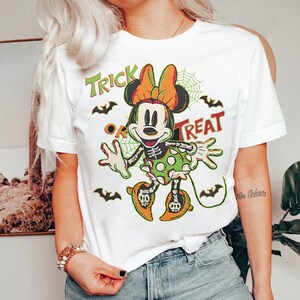 May include: White t-shirt featuring a Halloween-themed Minnie Mouse design. The graphic includes a skeleton Minnie, bats, spiderwebs, and the words "Trick or Treat" in green and orange. A fun and festive design.