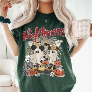 May include: Green t-shirt featuring a graphic print of Mickey Mouse and Minnie Mouse dressed as ghosts. The text "Nightmare on Main Street" is printed on the shirt.