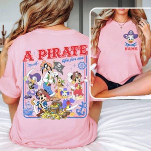 May include: Pink t-shirt featuring a pirate-themed design with cartoon characters. The back of the shirt has the text "A PIRATE life for me" with pirate-themed illustrations. The front of the shirt has a pirate Donald Duck design with the word "NAME" below.