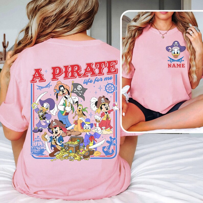May include: Pink t-shirt featuring a pirate-themed design with cartoon characters. The back of the shirt has the text "A PIRATE life for me" with pirate-themed illustrations. The front of the shirt has a pirate Donald Duck design with the word "NAME" below.