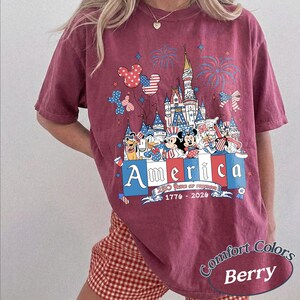May include: A berry-colored short-sleeve t-shirt featuring a graphic of a castle, fireworks, and cartoon characters wearing patriotic red, white, and blue clothing. The text reads America, 250 Years of Freedom, and 1776-2026 with balloon accents.