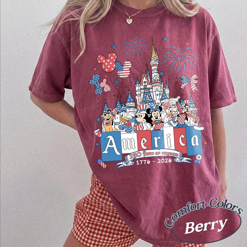 May include: A berry-colored short-sleeve t-shirt featuring a graphic of a castle, fireworks, and cartoon characters wearing patriotic red, white, and blue clothing. The text reads America, 250 Years of Freedom, and 1776-2026 with balloon accents.