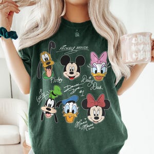 May include: A green t-shirt with a graphic print of Mickey Mouse, Minnie Mouse, Donald Duck, Daisy Duck, Goofy, and Pluto. The shirt has the text "Mickey Mouse", "Pluto", "Donald Duck #1", "Daisy Duck", "Goofy", and "Minnie Mouse XOXO" written on it.