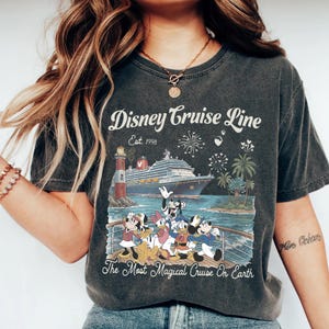 May include: Dark grey t-shirt with a Disney Cruise Line graphic. The design shows a cruise ship, lighthouse, fireworks, palm trees, and Disney characters. Text reads "Disney Cruise Line Est. 1998" and "The Most Magical Cruise On Earth."