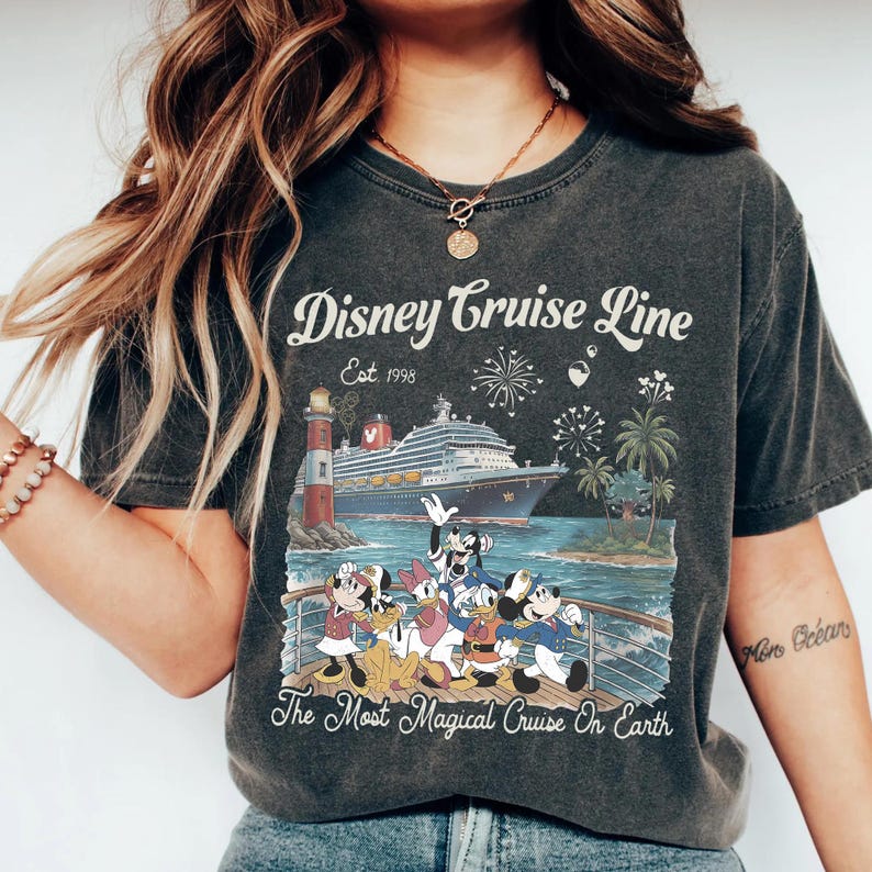 May include: Dark grey t-shirt with a Disney Cruise Line graphic. The design shows a cruise ship, lighthouse, fireworks, palm trees, and Disney characters. Text reads "Disney Cruise Line Est. 1998" and "The Most Magical Cruise On Earth."