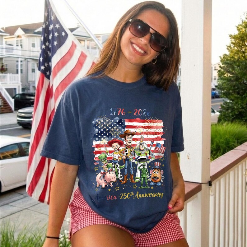 May include: A navy blue short-sleeve t-shirt featuring a graphic of Toy Story characters in front of an American flag with fireworks. The text reads 1776-2026 and America 250th Anniversary. The design includes Woody, Buzz Lightyear, Jessie, Rex, Hamm, and Mr. Potato Head.