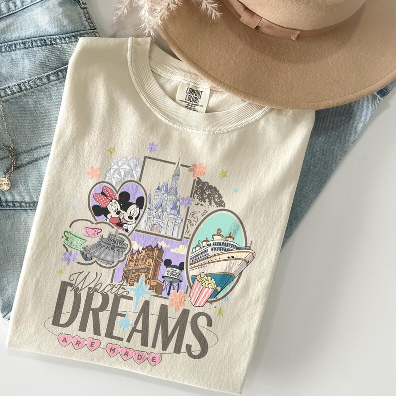 May include: Cream-colored t-shirt with a graphic design featuring Mickey and Minnie Mouse, a castle, a cruise ship, and the text "What Dreams Are Made Of." The shirt is folded on top of blue jeans.