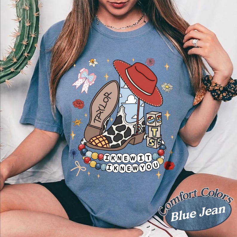 May include: A blue denim-coloured t-shirt featuring a graphic print of a cow-print cowboy boot, a red cowboy hat, a stack of blocks, and a colourful beaded necklace. The text "TAYLOR", "13", and "I KNEW IT I KNEW YOU" is printed on the shirt.