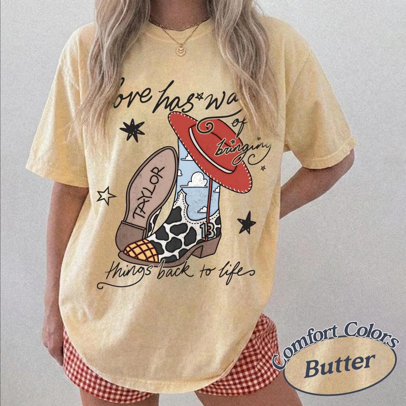 May include: A pale yellow short-sleeve t-shirt featuring a graphic of a cow-print cowboy boot, a red hat, and stars. The text reads, "Love has a way of bringing things back to life," with "TAYLOR" on the boot sole and the number "13" on the side.