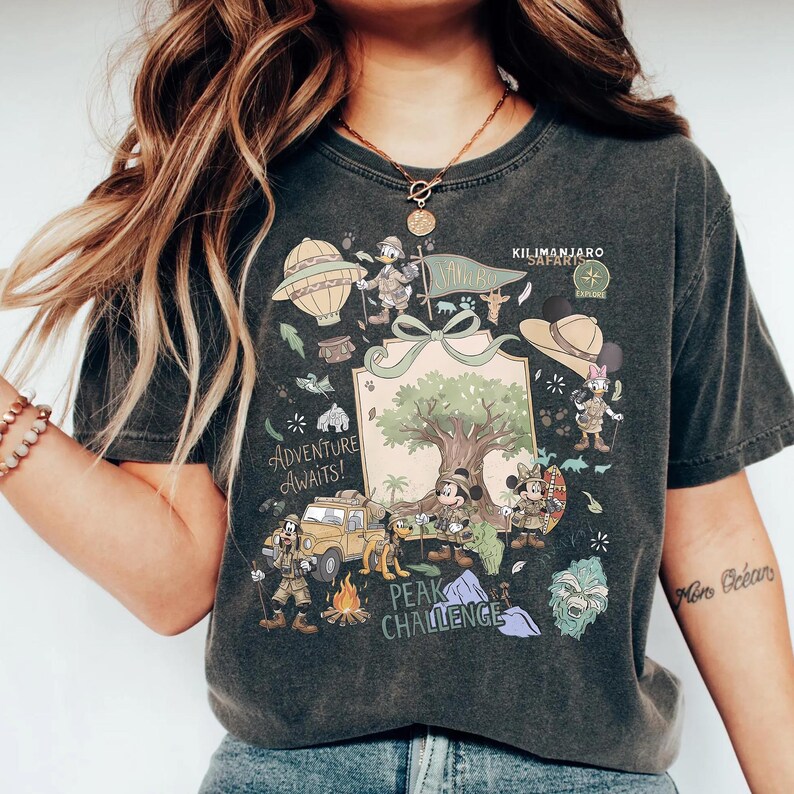 May include: Dark gray t-shirt featuring a colorful graphic of cartoon characters, a hot air balloon, and a safari jeep. The shirt has the words "Adventure Awaits!" and "Peak Challenge" printed on it. The shirt also has the words "Kilimanjaro Safari Explore" printed on it.