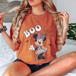 May include: Orange t-shirt with a Halloween-themed graphic. The design features the word "BOO" above a cartoon Minnie Mouse holding a pumpkin. A spiderweb and bats complete the spooky design.
