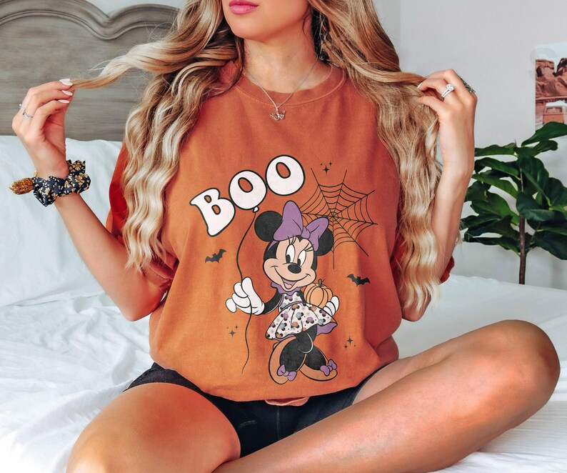 May include: Orange t-shirt with a Halloween-themed graphic. The design features the word "BOO" above a cartoon Minnie Mouse holding a pumpkin. A spiderweb and bats complete the spooky design.