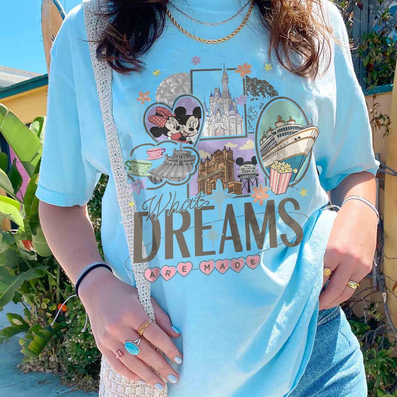 May include: Light blue t-shirt with a colorful graphic design featuring Mickey and Minnie Mouse, a castle, and a cruise ship. The text "What Dreams Are Made" is printed on the front. The shirt is a short-sleeved, casual style.