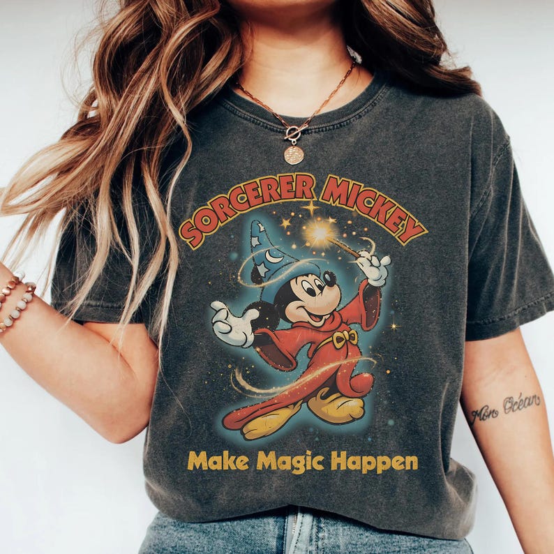 Peut inclure: T-shirt gris fonc&eacute; avec Mickey Mouse en sorcier de Disney. Mickey est repr&eacute;sent&eacute; dans sa tenue de sorcier embl&eacute;matique, tenant une baguette magique. Le texte "Sorcerer Mickey" est au-dessus de l'image, avec "Make Magic Happen" en dessous. Le t-shirt a un aspect vintage.