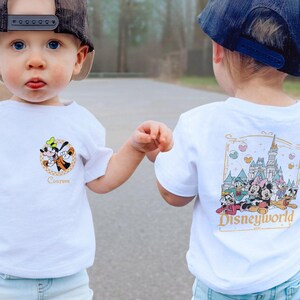 May include: Two toddlers wearing white t-shirts with Disney-themed graphics. One shirt features a cartoon image of Goofy with the word "Custom" below. The other shirt features a cartoon image of Cinderella's castle with Mickey Mouse, Minnie Mouse, Donald Duck, Goofy, Pluto, and other Disney characters. The text "Disneyworld 1971" is below the image.
