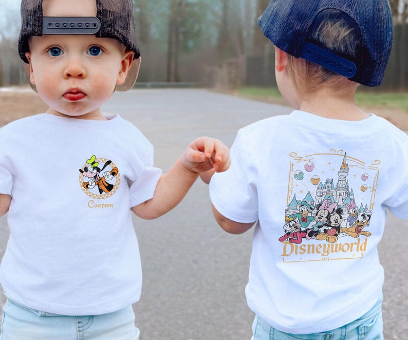 May include: Two toddlers wearing white t-shirts with Disney-themed graphics. One shirt features a cartoon image of Goofy with the word "Custom" below. The other shirt features a cartoon image of Cinderella's castle with Mickey Mouse, Minnie Mouse, Donald Duck, Goofy, Pluto, and other Disney characters. The text "Disneyworld 1971" is below the image.