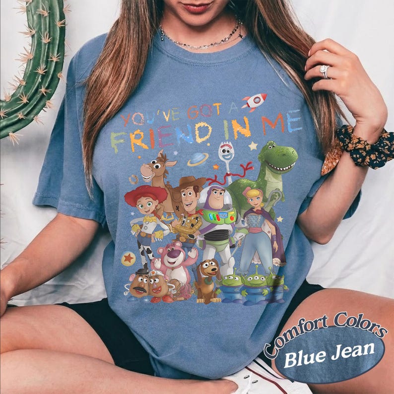 May include: A blue jean colored t-shirt featuring a graphic print of various animated characters from the Toy Story franchise. The text "YOU'VE GOT A FRIEND IN ME" is printed in colorful letters above the characters, which include Woody, Buzz Lightyear, Jessie, Rex, and others.