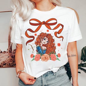May include: White t-shirt with a red bow and a cartoon illustration of a red-haired woman with a bow and arrow. The woman is surrounded by pink and orange flowers.