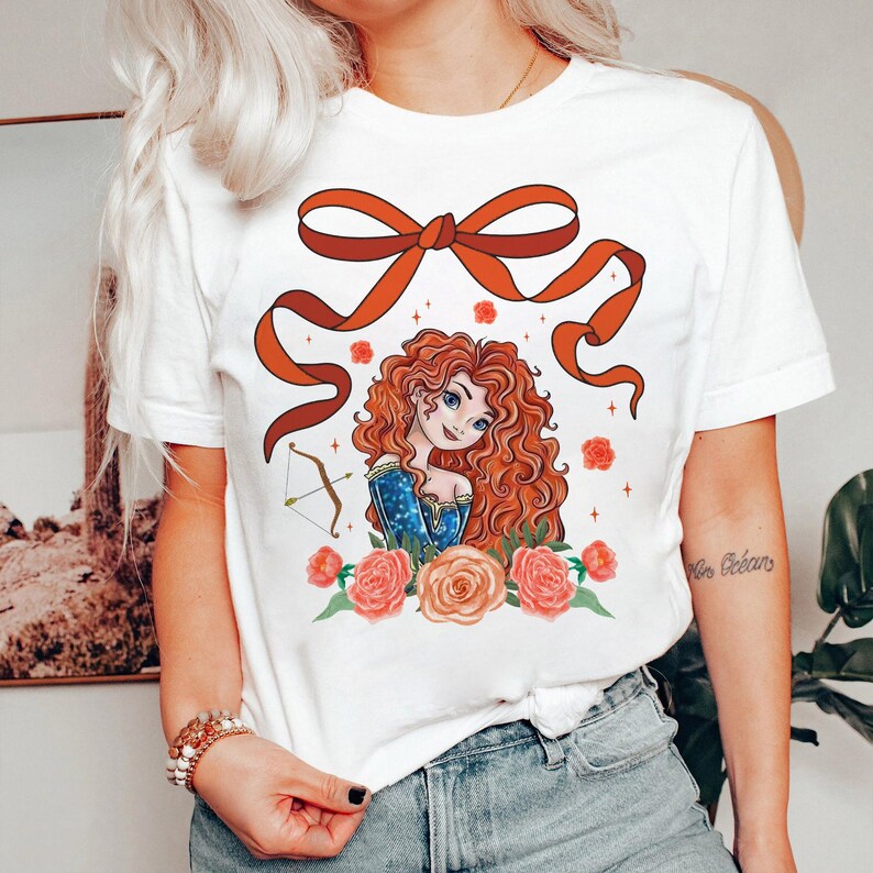 May include: White t-shirt with a red bow and a cartoon illustration of a red-haired woman with a bow and arrow. The woman is surrounded by pink and orange flowers.