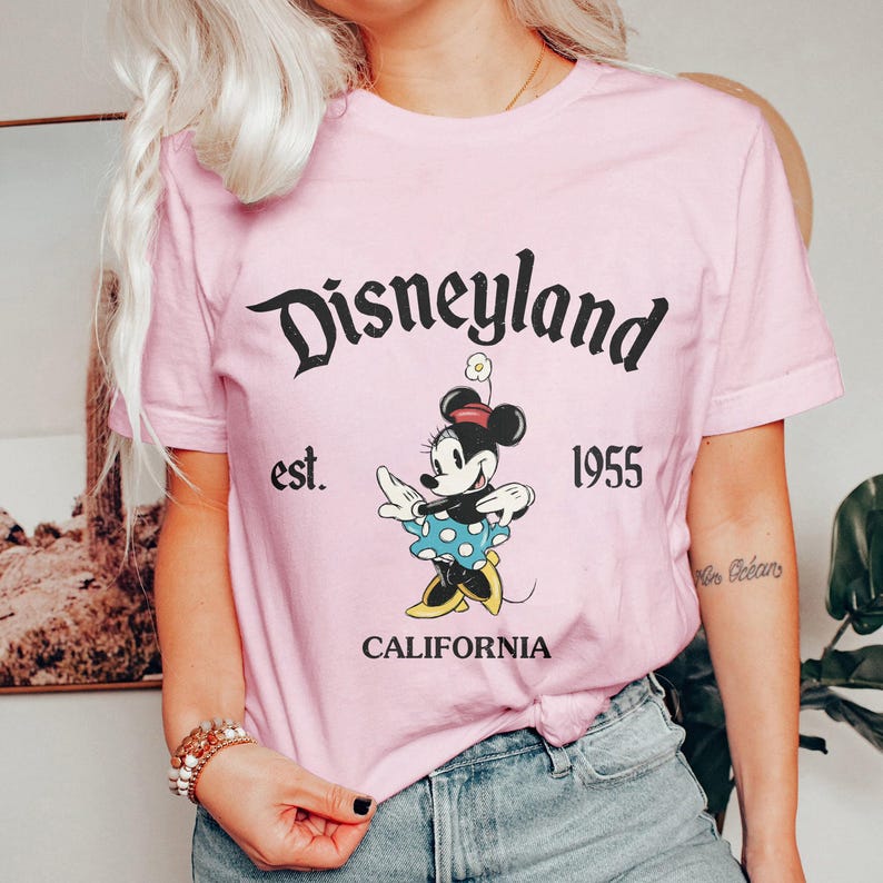 以下が含まれることがあります： ミッキーマウスの黒いグラフィックと「Disneyland est. 1955 CALIFORNIA」のテキストが書かれたピンクのTシャツ