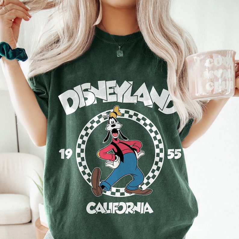 K&ouml;nnte beinhalten: Dunkelgr&uuml;nes T-Shirt mit dem Wort "Disneyland" in Wei&szlig; und einem Cartoon-Bild von Goofy. Das Shirt enth&auml;lt auch die Zahlen "19" und "55" und das Wort "California".