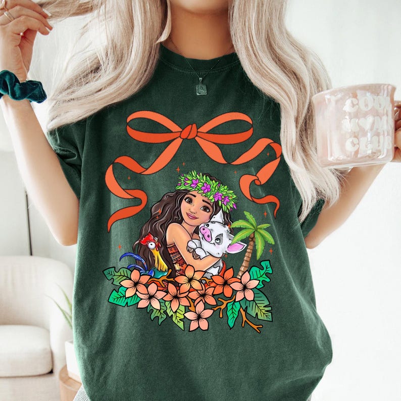 May include: A green t-shirt with a cartoon image of a Polynesian woman with a pig and a rooster. The woman is wearing a flower crown and holding the pig. The image is surrounded by flowers and palm trees. There is a large orange bow on the shirt.