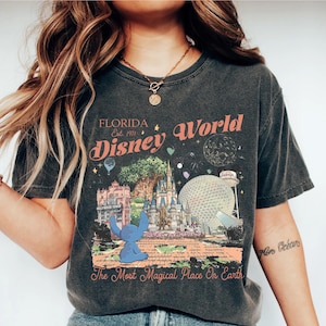 May include: Dark gray t-shirt featuring a colorful graphic of Disney World in Florida. The design includes the text "Florida Disney World Est. 1971" and "The Most Magical Place On Earth". A blue cartoon character is also featured.