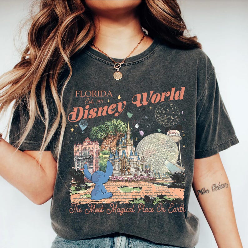 May include: Dark gray t-shirt featuring a colorful graphic of Disney World in Florida. The design includes the text "Florida Disney World Est. 1971" and "The Most Magical Place On Earth". A blue cartoon character is also featured.