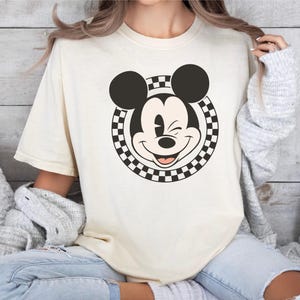 以下が含まれることがあります： 笑顔のミッキーマウスのグラフィックが特徴のクリーム色のTシャツ。グラフィックは、市松模様の円で囲まれたミッキーの顔の白黒画像です。シャツは半袖で、ゆったりとしたフィット感です。
