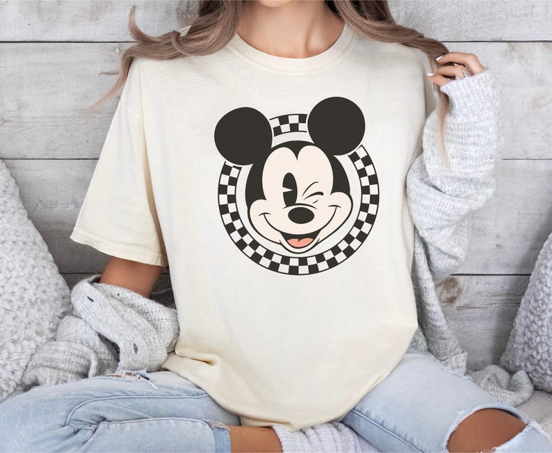 以下が含まれることがあります： 笑顔のミッキーマウスのグラフィックが特徴のクリーム色のTシャツ。グラフィックは、市松模様の円で囲まれたミッキーの顔の白黒画像です。シャツは半袖で、ゆったりとしたフィット感です。