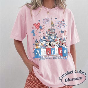 May include: A light pink t-shirt featuring a graphic of a castle, fireworks, and cartoon characters wearing patriotic red, white, and blue clothing. The text reads America, 250 Years of Freedom, and 1776-2026. Mickey-shaped balloons are also visible.