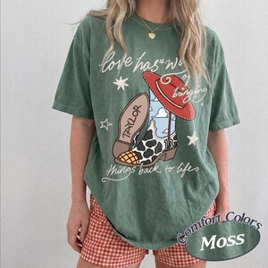 May include: A moss green short-sleeve t-shirt featuring a graphic of a red cowboy hat, a cow-print boot with the name Taylor and the number 13, and the text Love has a way of bringing things back to life, accented with white stars.