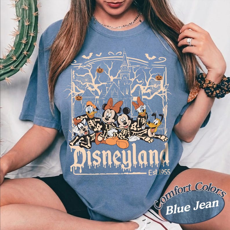 May include: Blue jean-colored t-shirt featuring a Halloween-themed Disneyland graphic. The design includes cartoon characters dressed as skeletons, a haunted castle, bats, and pumpkins. The text "Disneyland" and "Est. 1955" are also visible.