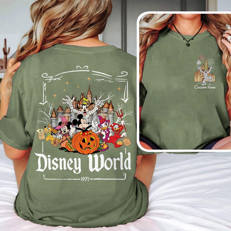 May include: Olive green t-shirt featuring a Disney World Halloween design. The graphic includes Mickey Mouse, Donald Duck, and other characters with a pumpkin and castle. The text "Disney World" and "1971" are also visible.