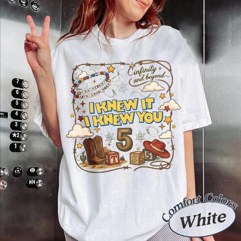 May include: A white t-shirt featuring a graphic print with a lasso border, the text "Cinfinity and beyond", "I KNEW IT I KNEW YOU", and "YOU'VE GOT A FRIEND IN ME". The design includes a cowboy hat, boots, blocks with letters, and the number 5.