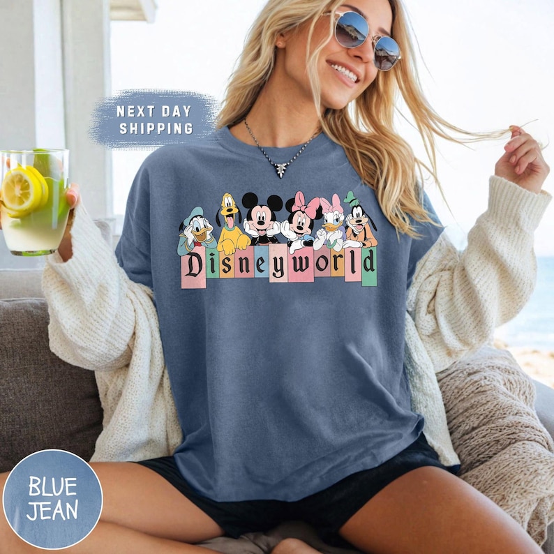 Pode incluir: Uma camiseta de jeans azul com um gr&aacute;fico de Mickey Mouse e seus amigos na frente de um letreiro colorido "Disneyworld".