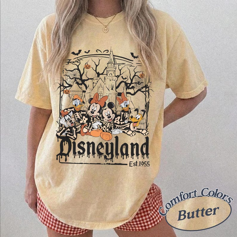 May include: Butter-colored t-shirt featuring a Halloween-themed Disneyland graphic. The design includes Mickey and Minnie Mouse, Donald and Daisy Duck, and Pluto in skeleton costumes, with a haunted castle backdrop. The text "Disneyland Est. 1955" is below the graphic.