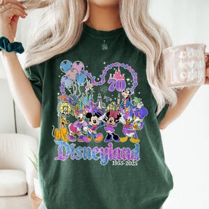 May include: Dark green t-shirt featuring a colorful Disneyland graphic. The design includes various Disney characters, balloons, a castle, and the text "Disneyland 1955-2025" celebrating its 70th anniversary. The shirt is a comfortable, casual style.