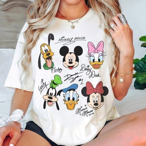 May include: White t-shirt with a graphic print of Mickey Mouse, Minnie Mouse, Donald Duck, Daisy Duck, Goofy, and Pluto. The characters are drawn in a cartoon style and have their names written underneath them. The text "Mickey Mouse", "Donald Duck #1", "Daisy Duck", "Pluto", "Goofy", and "Minnie Mouse xoxo" is written on the shirt.