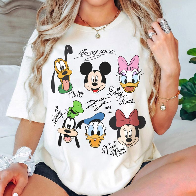 May include: White t-shirt with a graphic print of Mickey Mouse, Minnie Mouse, Donald Duck, Daisy Duck, Goofy, and Pluto. The characters are drawn in a cartoon style and have their names written underneath them. The text "Mickey Mouse", "Donald Duck #1", "Daisy Duck", "Pluto", "Goofy", and "Minnie Mouse xoxo" is written on the shirt.