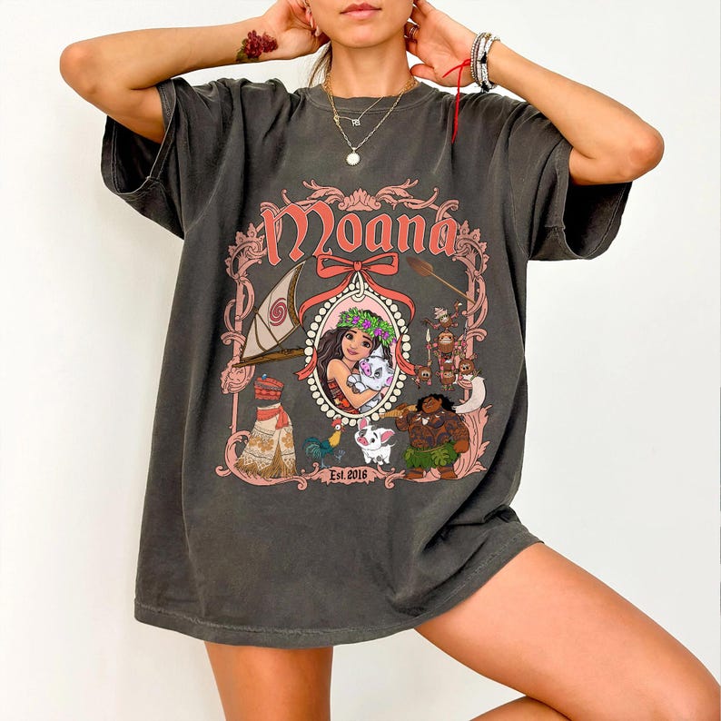 Puede incluir: Una camiseta gris oscuro de gran tama&ntilde;o con un estampado gr&aacute;fico de Moana y personajes de la pel&iacute;cula. El gr&aacute;fico incluye el texto "Moana" y "Est. 2016".