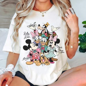 May include: Cream-colored t-shirt featuring a colorful graphic of Disney characters including Mickey Mouse, Minnie Mouse, Donald Duck, Daisy Duck, Goofy, and Pluto. The characters' names are written in cursive. The shirt is a casual style.