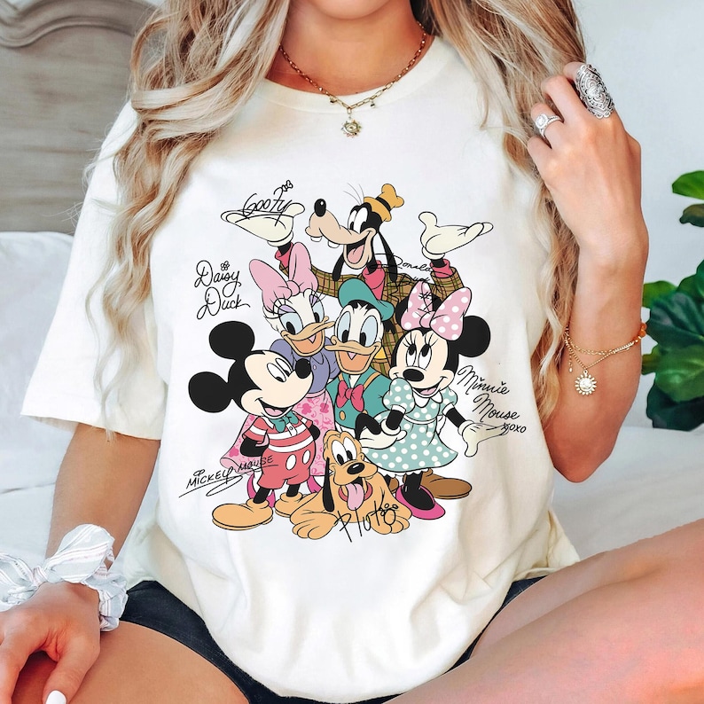 May include: Cream-colored t-shirt featuring a colorful graphic of Disney characters including Mickey Mouse, Minnie Mouse, Donald Duck, Daisy Duck, Goofy, and Pluto. The characters' names are written in cursive. The shirt is a casual style.