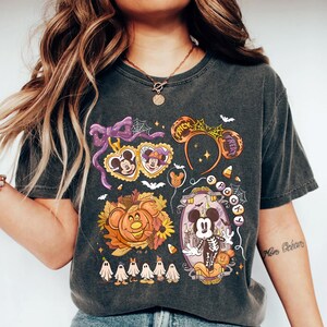 May include: Dark gray t-shirt featuring a colorful Halloween-themed graphic. The design includes Mickey Mouse in a skeleton costume, a pumpkin, ghosts, and the words "Trick or Treat" on a headband. The shirt has a vintage wash.