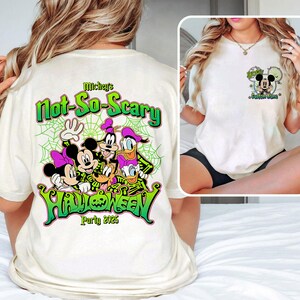 Op de afbeelding: Cr&egrave;mekleurig T-shirt met een Halloween-ontwerp met Mickey Mouse, Minnie Mouse, Goofy, Donald Duck en Daisy Duck. Het shirt heeft de tekst "Mickey's Not-So-Scary Halloween Party 2025" in groen en zwart.