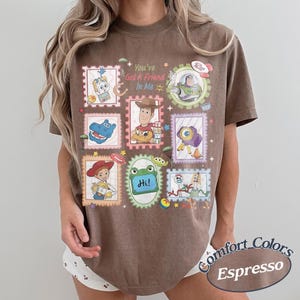 May include: A brown short-sleeve t-shirt featuring a graphic print of various Toy Story characters arranged in postage stamp-style frames. The text "You've Got A Friend In Me" is printed in the center, surrounded by illustrations of Woody, Buzz Lightyear, Jessie, and other characters.